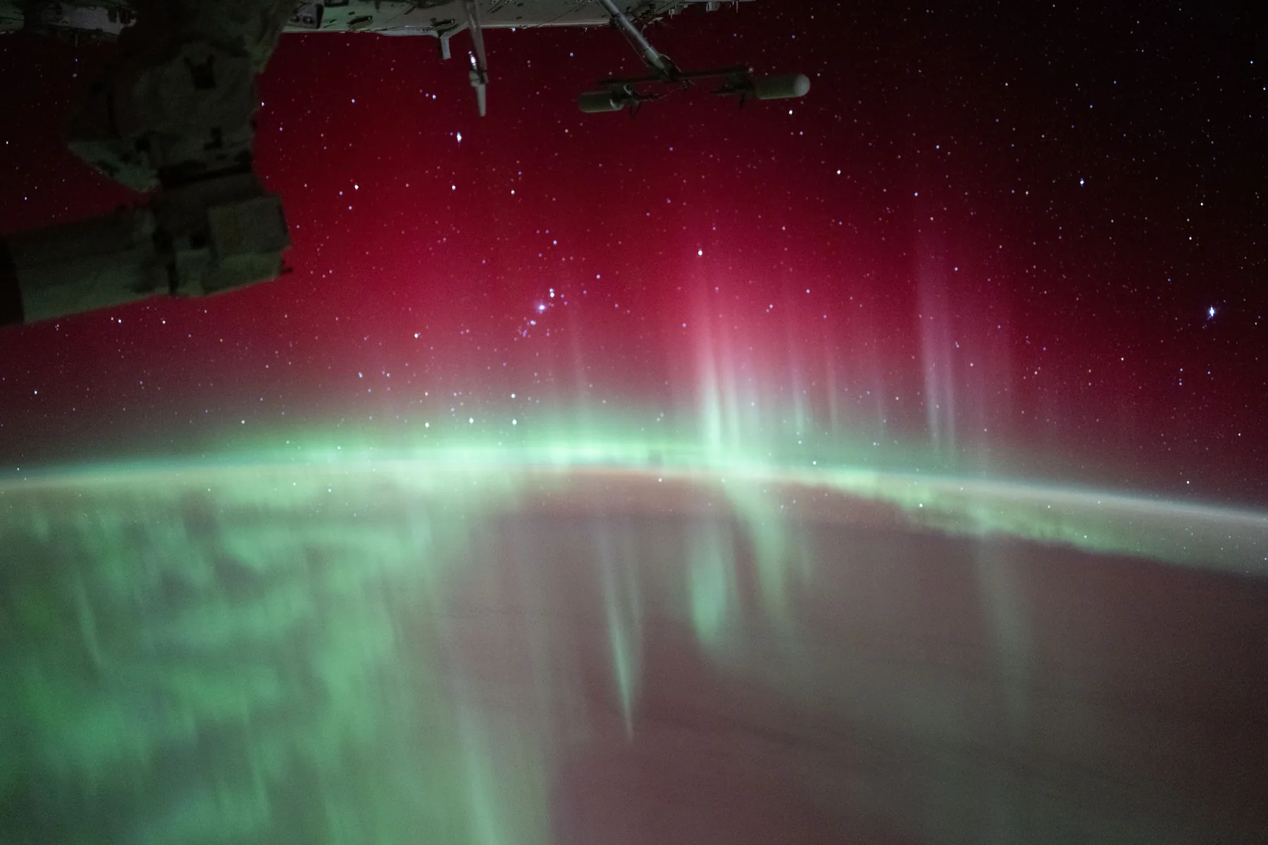 Green and red aurora curtains stretch over Earth’s curved night horizon, with stars beyond and part of the space station overhead.